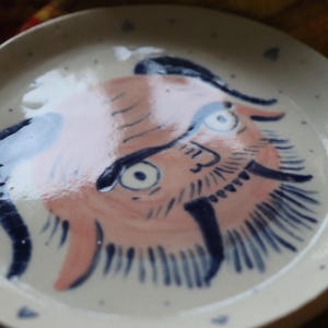May include: A white ceramic plate with a hand-painted design of a pink and blue creature with a beard and horns. The creature has a whimsical expression and is painted in a folk art style.