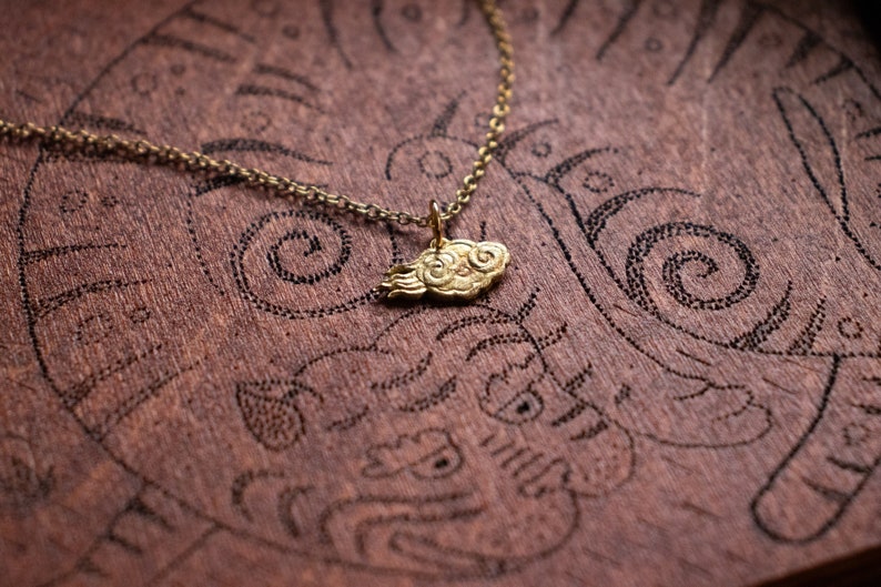 May include: A gold necklace with a cloud-shaped pendant. The pendant is detailed with swirls and is on a brown wooden surface.