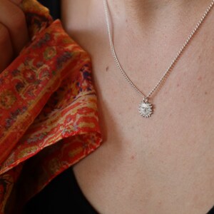 May include: A silver necklace with a small sun pendant. The pendant has a detailed design with a face in the center.