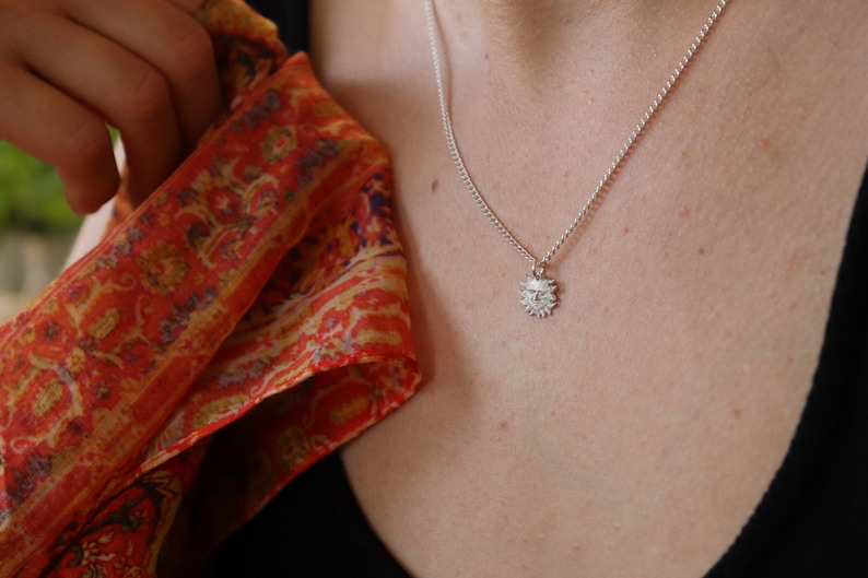 May include: A silver necklace with a small sun pendant. The pendant has a detailed design with a face in the center.