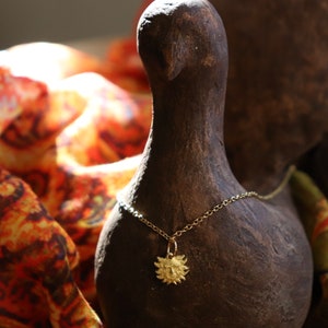 May include: A gold necklace with a sun pendant hangs on a brown ceramic bird figurine. The bird is surrounded by a colorful patterned fabric.