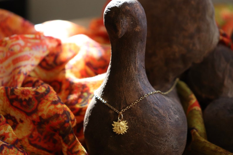 May include: A gold necklace with a sun pendant hangs on a brown ceramic bird figurine. The bird is surrounded by a colorful patterned fabric.