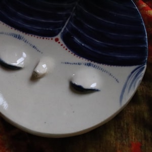 May include: A white ceramic dish with a blue and red design. The dish features a stylised face with closed eyes and a small nose.