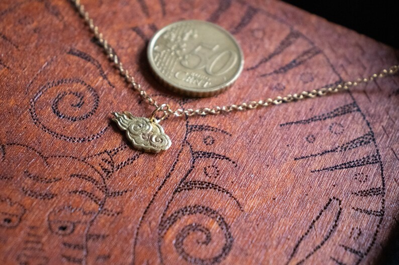 May include: A gold chain necklace with a cloud-shaped pendant. The necklace is lying on a wooden surface with a 50 cent coin.