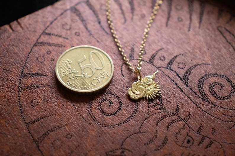 Op de afbeelding: Een gouden ketting met een bedel die een zon en wolken voorstelt. De bedel ligt op een houten oppervlak met een munt van 50 eurocent.