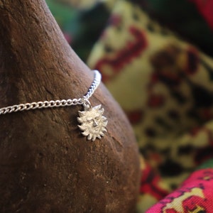 May include: A silver chain necklace with a sun face pendant. The pendant is detailed with a face and rays. The necklace is on a brown textured surface.