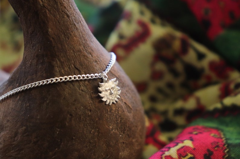 May include: A silver chain necklace with a sun face pendant. The pendant is detailed with a face and rays. The necklace is on a brown textured surface.