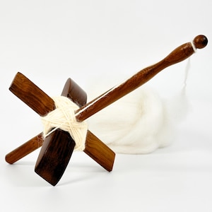 May include: A wooden drop spindle with a cross-shaped whorl and a long, tapered shaft. The spindle is made of dark wood and has a ball at the end of the shaft. White yarn is wrapped around the whorl, and a fluffy cloud of white wool sits nearby.