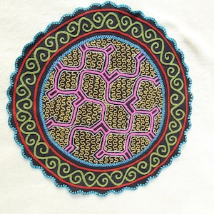 Op de afbeelding: Cirkelvormig, geborduurd textiel met een zwarte achtergrond. Het ontwerp heeft een centraal patroon van roze en gele geometrische vormen, omgeven door concentrische ringen van rood, blauw en groen. De buitenrand heeft een geschulpte rand.
