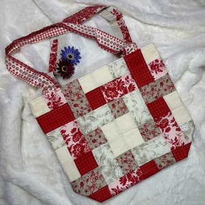 May include: A patchwork tote bag with a white background and red, pink, and beige floral patterns. The bag has two straps and is made of fabric.