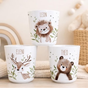 Könnte beinhalten: Drei weiße zylindrische Becher mit Illustrationen eines Löwen, eines Rehs und eines Bären. Jeder Becher ist mit einem Namen personalisiert: Leon über dem Löwen, Eleni über dem Reh und Theo über dem Bären, verziert mit grünen Blättern.