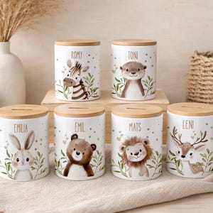 Könnte beinhalten: Sechs zylindrische Spardosen aus weißer Keramik mit hellen Holzdeckeln, jede mit einer individuellen Aquarell-Tierillustration und einem Namen: Romy das Zebra, Toni der Otter, Emilia der Hase, Emil der Bär, Mats der Löwe und Leni das Reh.