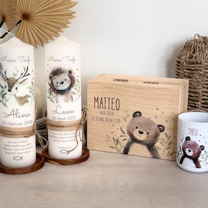Pu&ograve; includere: Due candele cilindriche bianche con spago, ciascuna con un'illustrazione di animale, il testo Meine Taufe, un nome e una data. Una scatola rettangolare in legno con l'illustrazione di un orso e il nome Matteo, e una tazza bianca con un orso e il nome Theo.