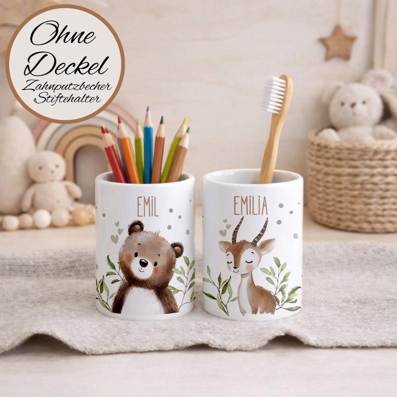 May include: Two white ceramic cylindrical cups featuring illustrations of a brown bear and a deer with green foliage. One cup is labeled Emil and holds colored pencils, while the other is labeled Emilia and holds a bamboo toothbrush.
