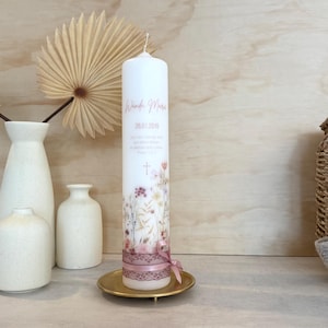 May include: A tall white cylindrical candle featuring a printed floral design in muted tones, the name Wanda Marie, the date 26.01.2019, and a small cross. The base is wrapped in a dusty pink lace ribbon tied into a small bow on a gold metal plate.