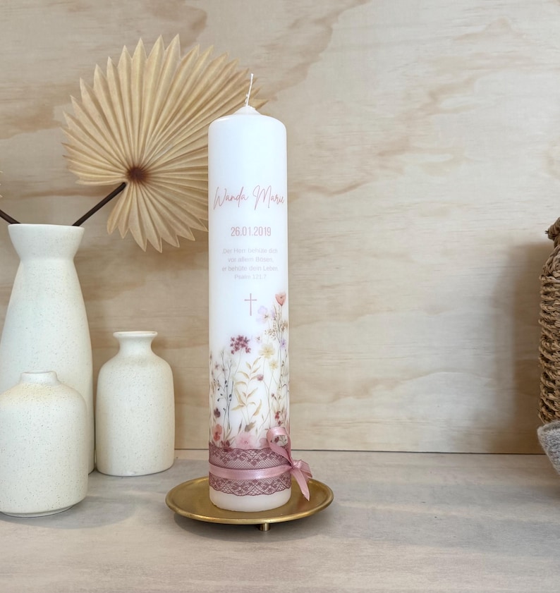 May include: A tall white cylindrical candle featuring a printed floral design in muted tones, the name Wanda Marie, the date 26.01.2019, and a small cross. The base is wrapped in a dusty pink lace ribbon tied into a small bow on a gold metal plate.