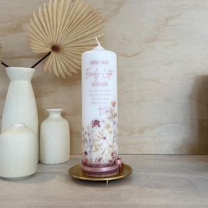 May include: A white cylindrical pillar candle featuring a floral print in muted tones, the text MEINE TAUFE, Emily Lotta, 29.03.2026, and a cross. The base is wrapped in a dusty rose ribbon with lace trim, resting on a small gold-colored metal plate.