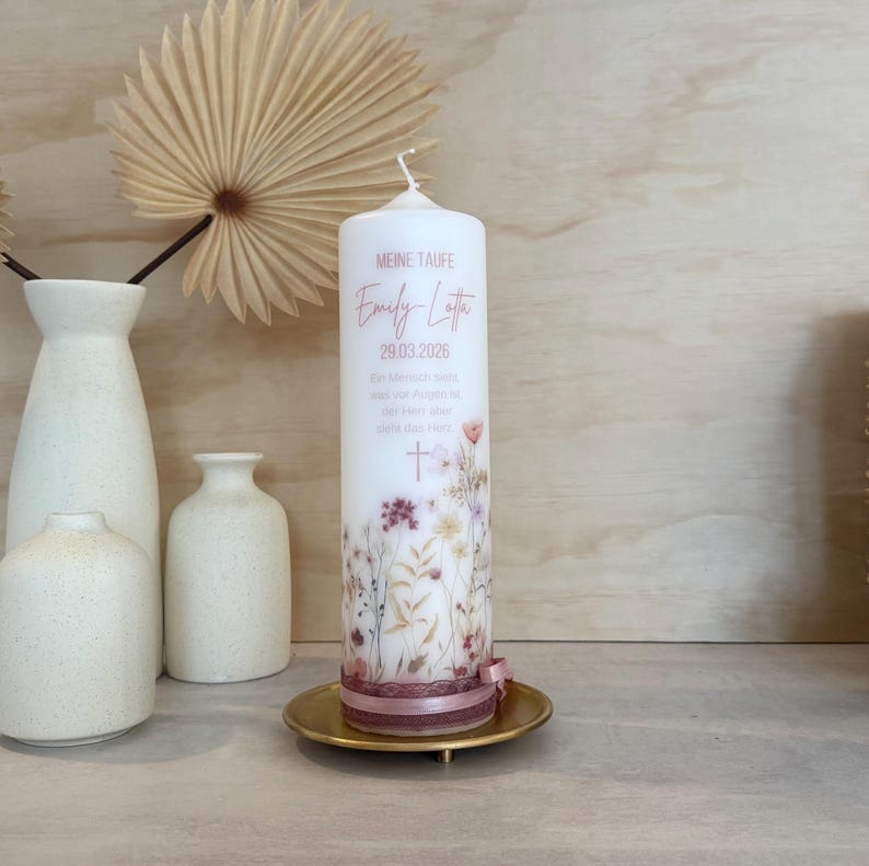 May include: A white cylindrical pillar candle featuring a floral print in muted tones, the text MEINE TAUFE, Emily Lotta, 29.03.2026, and a cross. The base is wrapped in a dusty rose ribbon with lace trim, resting on a small gold-colored metal plate.