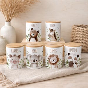 May include: Six cylindrical white ceramic money boxes with light wooden lids, each featuring a unique watercolor animal illustration and a name: Moritz, Finja, Hannes, Henri, James, and Matteo. The animals include a bear, horse, pig, koala, lion, and panda.
