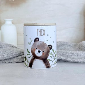 May include: A white cylindrical ceramic canister with a light wood lid. The canister features a printed illustration of a brown bear surrounded by green leaves, small hearts, and dots. The name THEO is printed in brown letters above the bear.