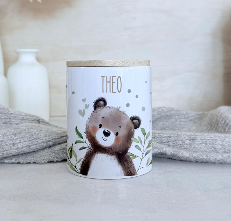 May include: A white cylindrical ceramic canister with a light wood lid. The canister features a printed illustration of a brown bear surrounded by green leaves, small hearts, and dots. The name THEO is printed in brown letters above the bear.