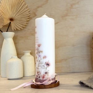 May include: A white pillar candle decorated with a delicate floral print featuring wildflowers in soft pink, purple, and green tones. The base is wrapped in a dark lace trim and a light pink ribbon, resting on a small, circular wooden stand.