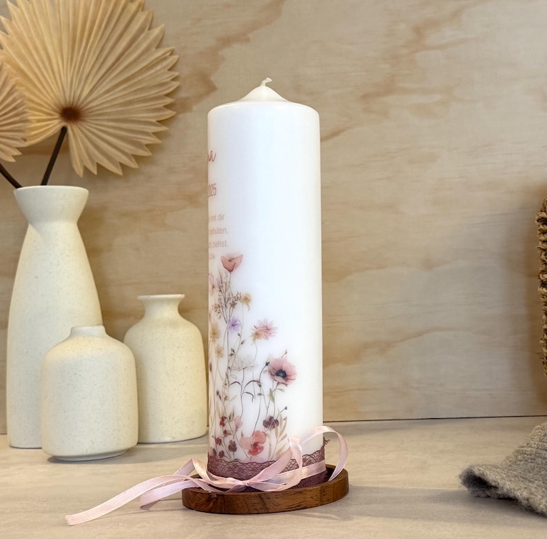 May include: A white pillar candle decorated with a delicate floral print featuring wildflowers in soft pink, purple, and green tones. The base is wrapped in a dark lace trim and a light pink ribbon, resting on a small, circular wooden stand.
