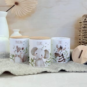 May include: Three white ceramic cylindrical coin banks with light wood lids. Each bank features a watercolor illustration of a baby animal: a squirrel labeled Paulina, an elephant labeled Elias, and a zebra labeled Zoe, surrounded by green leaves.