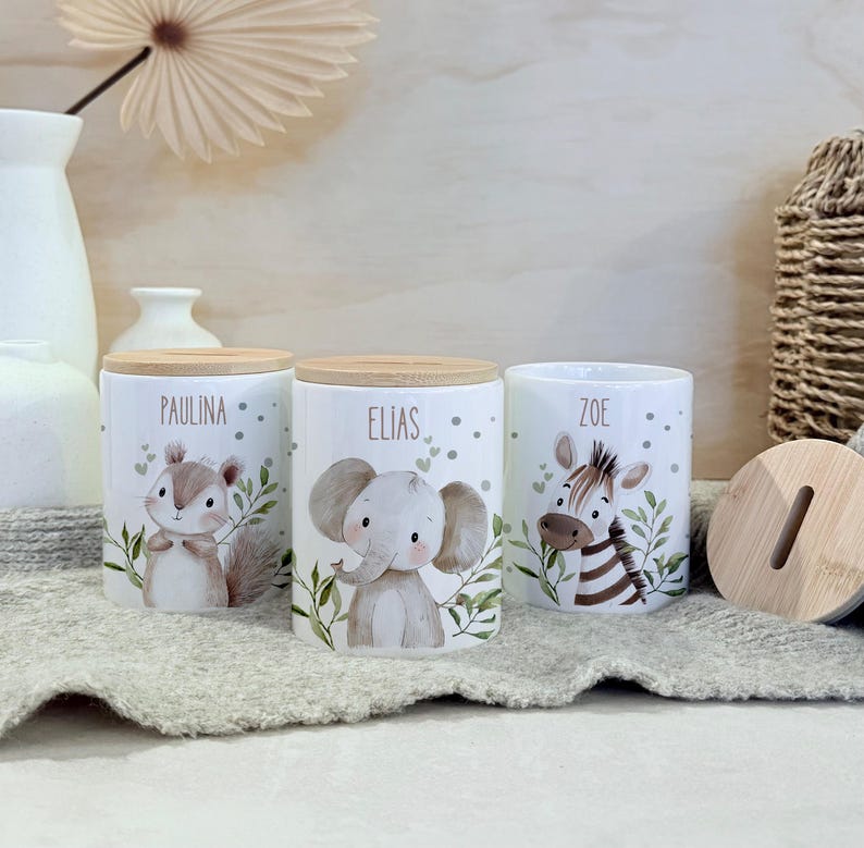 May include: Three white ceramic cylindrical coin banks with light wood lids. Each bank features a watercolor illustration of a baby animal: a squirrel labeled Paulina, an elephant labeled Elias, and a zebra labeled Zoe, surrounded by green leaves.