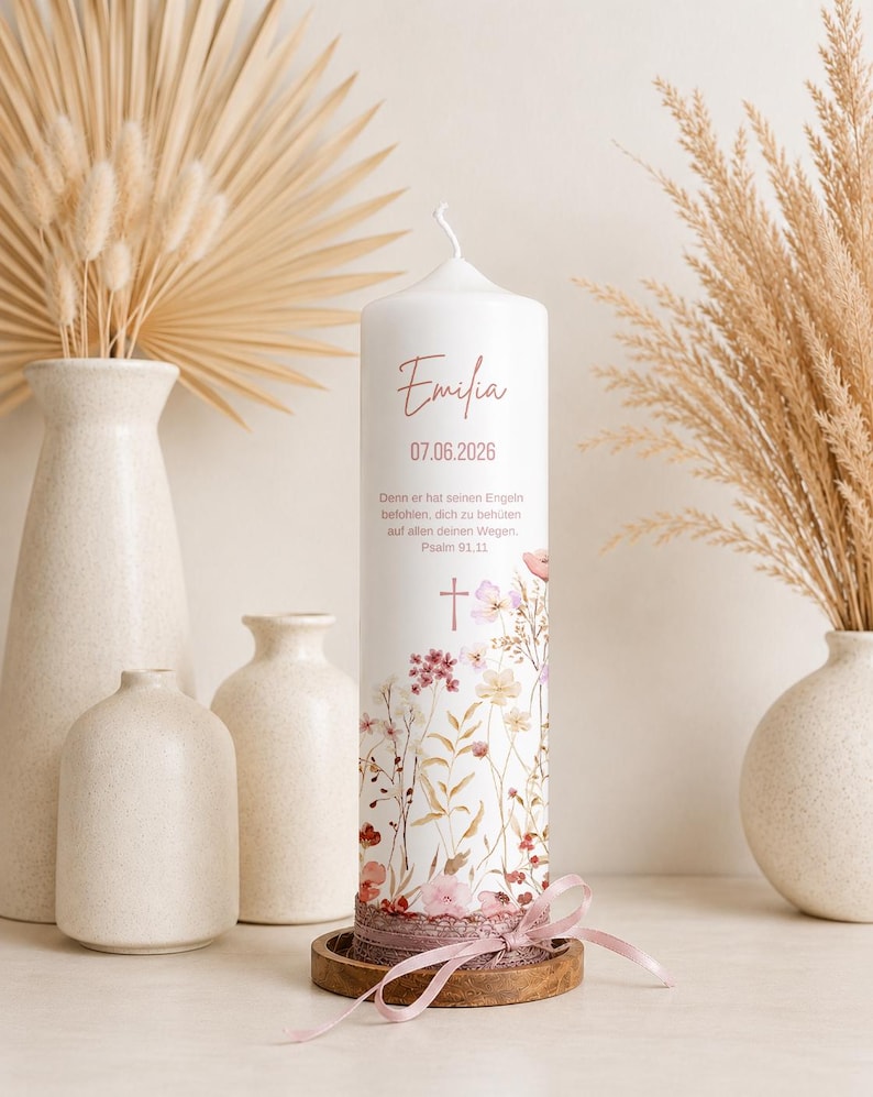 May include: A white cylindrical pillar candle featuring the name Emilia, the date 07.06.2026, and the German text Psalm 91.11. The candle is decorated with a floral print in muted pink and brown tones, a small cross, and a pink ribbon tied at the base.