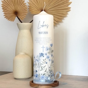 May include: A white cylindrical pillar candle featuring a blue floral print, a cross icon, and the text Lukas 19.07.2026. The candle includes a German Bible verse, a light blue ribbon tied at the base, and sits on a circular wooden stand.