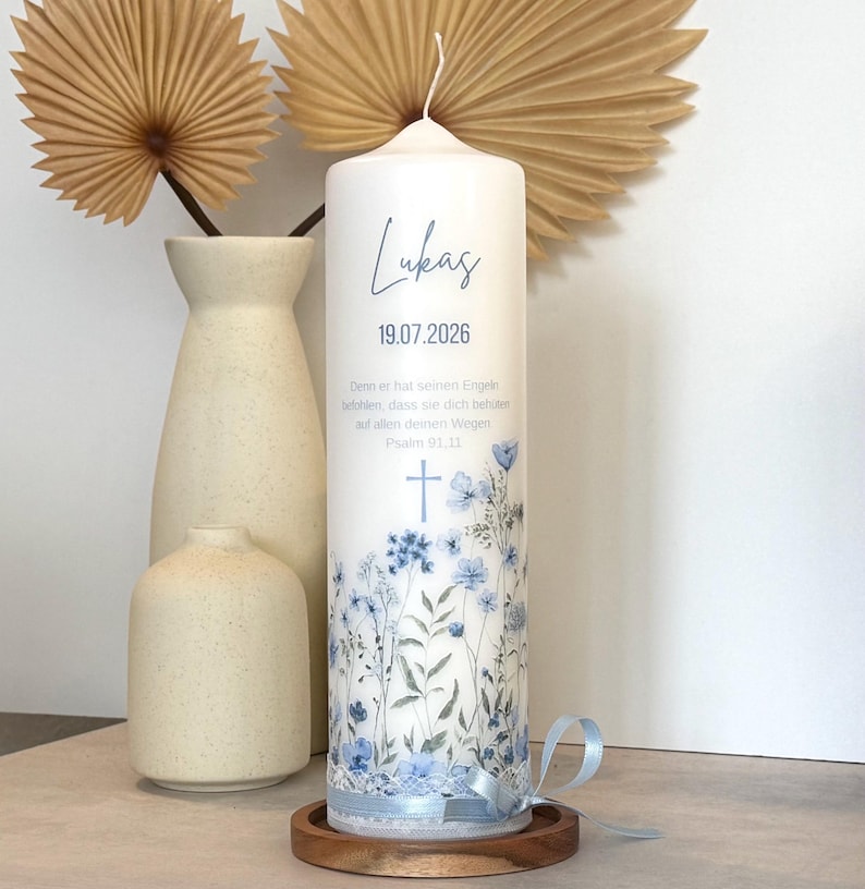 May include: A white cylindrical pillar candle featuring a blue floral print, a cross icon, and the text Lukas 19.07.2026. The candle includes a German Bible verse, a light blue ribbon tied at the base, and sits on a circular wooden stand.