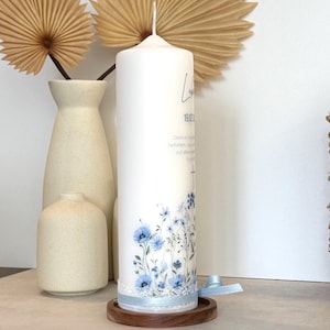 May include: A white cylindrical pillar candle featuring a blue floral print at the base, a light blue ribbon with white lace trim, and black printed text including a date and a cross symbol, resting on a circular wooden base.