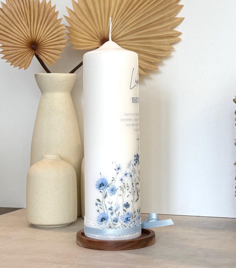 May include: A white cylindrical pillar candle featuring a blue floral print at the base, a light blue ribbon with white lace trim, and black printed text including a date and a cross symbol, resting on a circular wooden base.