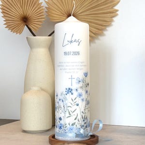 May include: A white cylindrical pillar candle featuring a blue floral print, a cross icon, and the text Lukas 19.07.2026, along with the German verse Psalm 91.11. The base is decorated with white lace trim and a light blue satin ribbon tied in a bow.