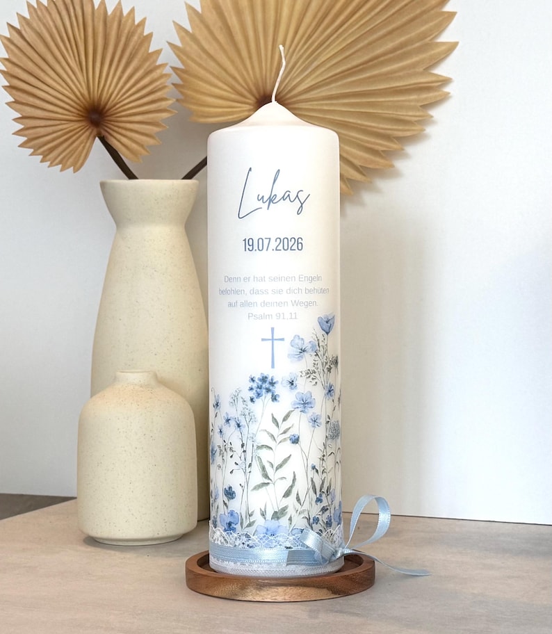 May include: A white cylindrical pillar candle featuring a blue floral print, a cross icon, and the text Lukas 19.07.2026, along with the German verse Psalm 91.11. The base is decorated with white lace trim and a light blue satin ribbon tied in a bow.