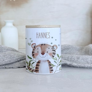 May include: A white cylindrical ceramic storage jar with a light wood lid. The jar features a watercolor illustration of a tiger cub surrounded by green leaves and small hearts. The name HANNES is printed in brown text above the tiger cub.