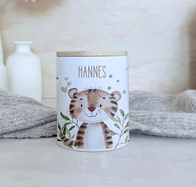 May include: A white cylindrical ceramic storage jar with a light wood lid. The jar features a watercolor illustration of a tiger cub surrounded by green leaves and small hearts. The name HANNES is printed in brown text above the tiger cub.