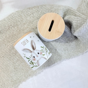 May include: A white cylindrical ceramic money box featuring a light brown wooden lid with a coin slot. The side of the container displays a printed illustration of a rabbit with green leaves and the name Sofia written in a dark grey sans-serif font.