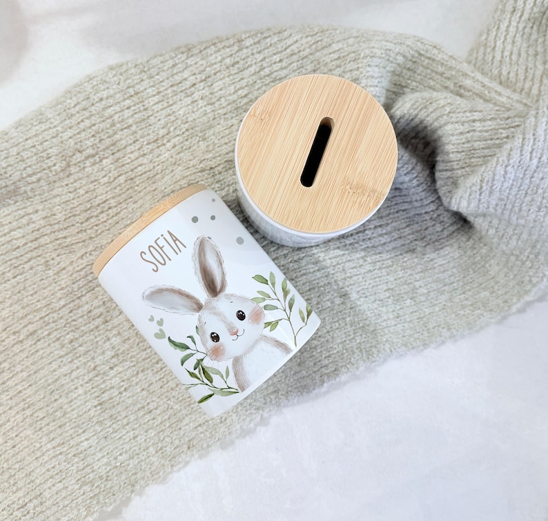 May include: A white cylindrical ceramic money box featuring a light brown wooden lid with a coin slot. The side of the container displays a printed illustration of a rabbit with green leaves and the name Sofia written in a dark grey sans-serif font.