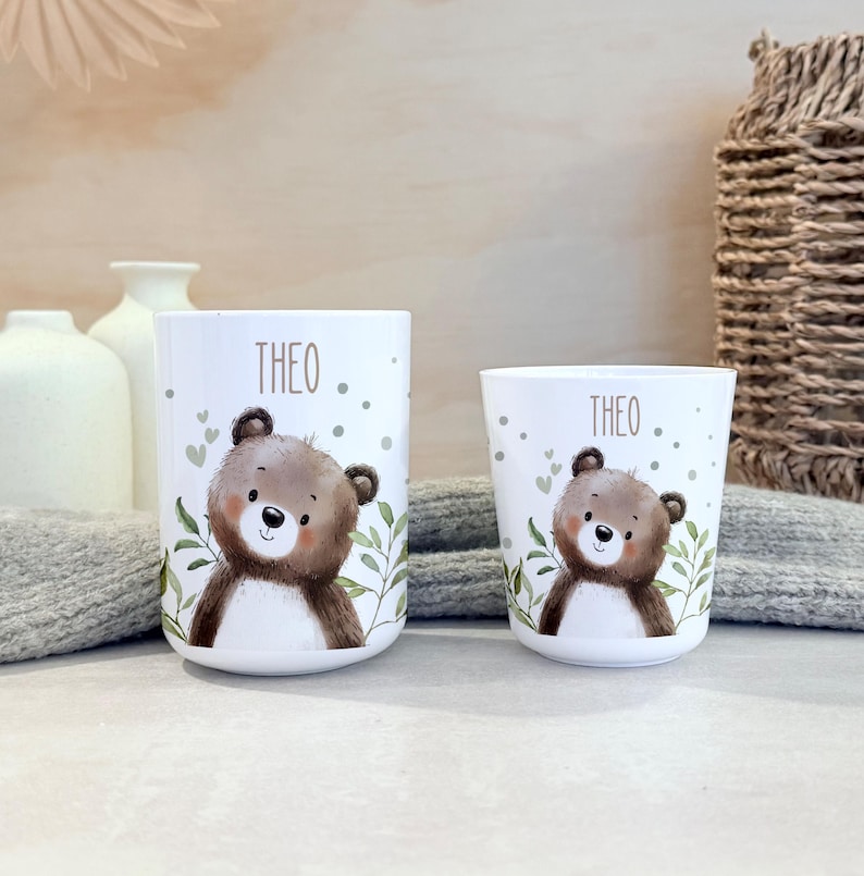 May include: Two white cylindrical cups of different sizes, each featuring a brown bear illustration, green leaf accents, and the name THEO printed in brown text above the bear. The cups have a smooth finish and a simple, rounded design.