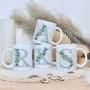 Könnte beinhalten: Weiße Keramikbecher mit großen Anfangsbuchstaben, verziert mit Eukalyptusgrün. Die Becher zeigen die Buchstaben "A", "R", "K" und "S" mit den Namen "Anna", "Rita", "Kiara" und "Svenja". Ein dekoratives Geschenk.