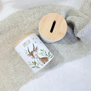May include: A white cylindrical ceramic money box featuring a light brown deer illustration, green leaves, and the name AMELIA printed in black. The container has a round, light-colored bamboo lid with a vertical coin slot for inserting money.