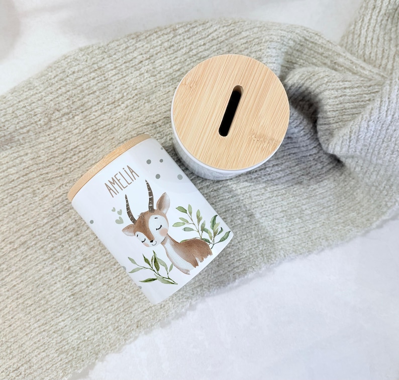May include: A white cylindrical ceramic money box featuring a light brown deer illustration, green leaves, and the name AMELIA printed in black. The container has a round, light-colored bamboo lid with a vertical coin slot for inserting money.