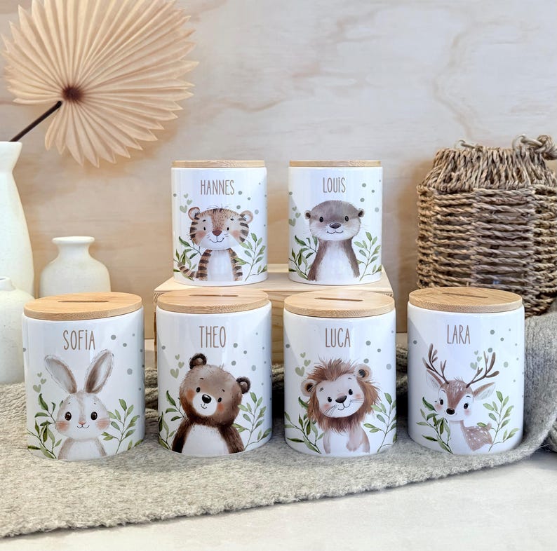 May include: Six white cylindrical ceramic money boxes with light wood lids, each featuring a unique watercolor animal illustration and a name: Hannes with a tiger, Louis with an otter, Sofia with a rabbit, Theo with a bear, Luca with a lion, and Lara with a deer.