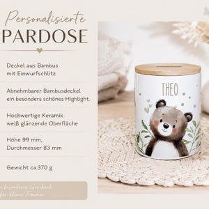 May include: A white ceramic money box with a glossy finish, featuring a brown bear illustration and the name THEO printed in black. It has a removable bamboo lid with a coin slot. The dimensions are 99 mm in height and 83 mm in diameter.