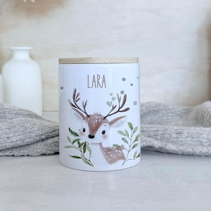 May include: A white ceramic cylindrical canister with a light wood lid. The canister features a printed illustration of a brown fawn surrounded by green leaves and small hearts. The name Lara is printed in brown text above the fawn's head.