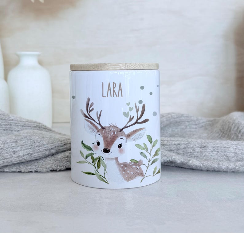 May include: A white ceramic cylindrical canister with a light wood lid. The canister features a printed illustration of a brown fawn surrounded by green leaves and small hearts. The name Lara is printed in brown text above the fawn's head.
