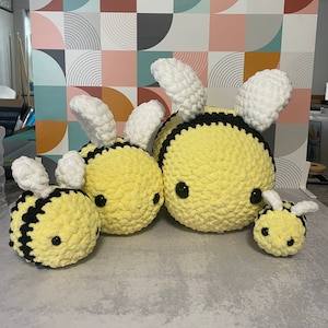 May include: Four yellow and black crochet bee plushies of varying sizes are arranged in a group. Each bee has white wings and black eyes. The bees are on a light gray surface with a colorful geometric patterned background.