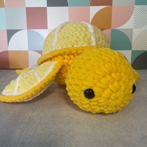May include: A crocheted yellow and white turtle toy with black eyes. The turtle is sitting on a light gray surface.
